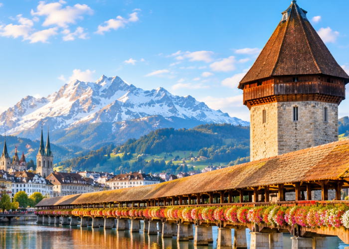 Chapel Bridge and Water Tower in Lucerne, Switzerland, with flower-lined railings, a swan on the river, and snow-capped mountains at sunrise.
