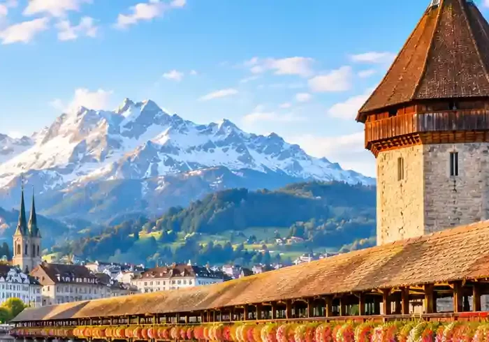 Chapel Bridge and Water Tower in Lucerne, Switzerland, with flower-lined railings, a swan on the river, and snow-capped mountains at sunrise.